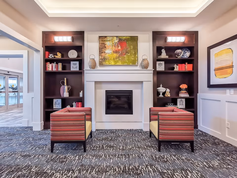 A cozy sitting area in a senior living facility featuring two red patterned armchairs facing a white fireplace with a colorful abstract painting above it. On either side of the fireplace are dark wooden built-in shelves decorated with books, vases, and decorative objects. The floor is carpeted with a patterned design, and there is a large windowed doorway to the left leading to another room.