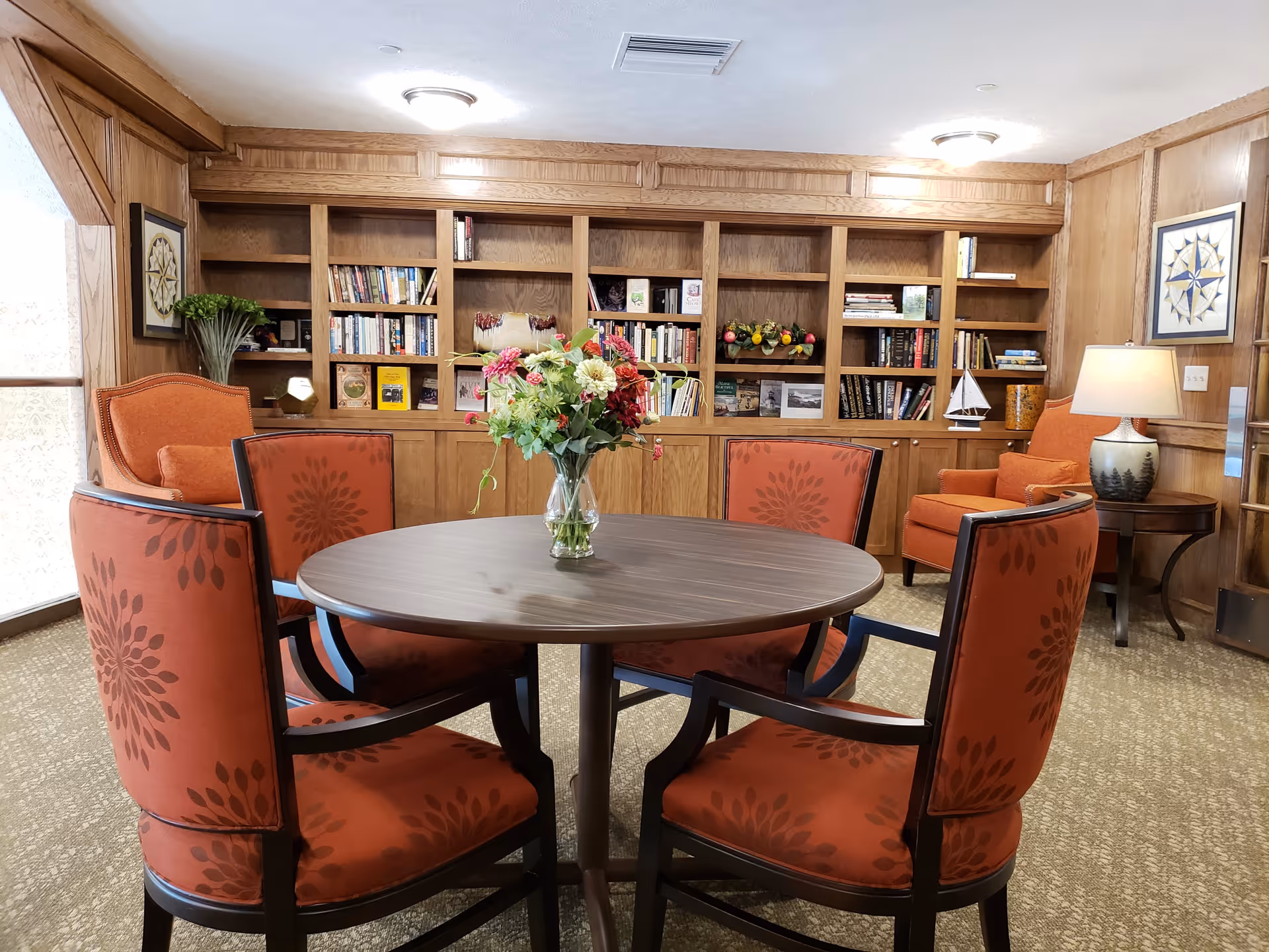 A cozy room with a round wooden table surrounded by four orange upholstered chairs with floral patterns. In the center of the table is a glass vase with a bouquet of flowers. Behind the table is a large wooden bookshelf filled with books and decorative items. Two orange armchairs are placed near the bookshelf, one next to a small side table with a lamp. The room has wood-paneled walls and carpeted flooring.