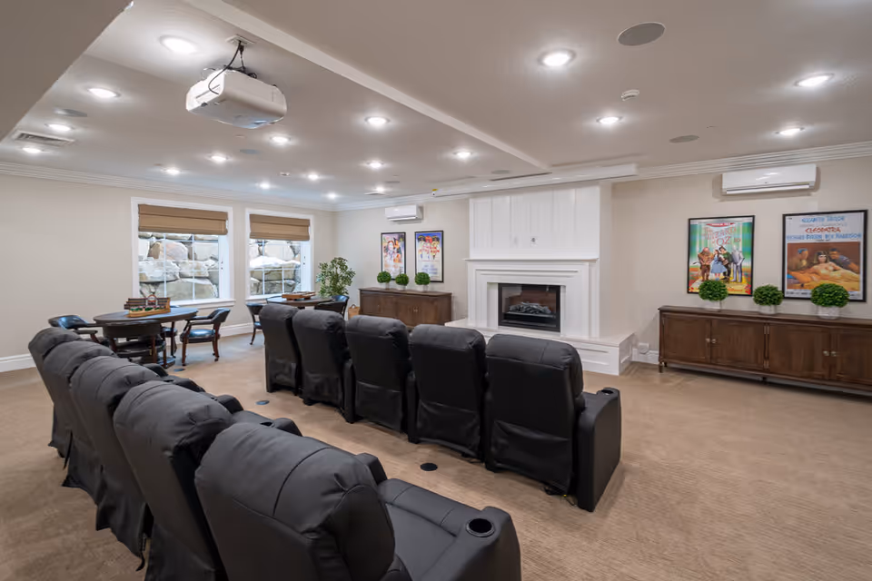 A cozy senior living common area with two rows of black recliner chairs facing a white fireplace. Above the chairs, a ceiling-mounted projector is visible. The room has beige walls, carpeted floor, and recessed lighting. There are two windows with brown blinds on the left side, two wooden cabinets with green potted plants and framed movie posters on the walls.