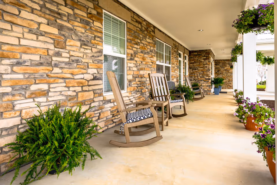 A covered outdoor porch area with stone walls, several wooden rocking chairs with patterned cushions, and various potted plants and hanging flower baskets along the edge.