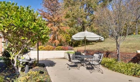 Outdoor patio area with a round table and four metal chairs, one of which has a cushion. A large striped umbrella provides shade over the table. The patio is surrounded by greenery including bushes, trees with autumn-colored leaves, and a grassy hill with a brick retaining wall.