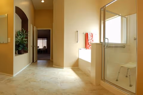 A bright bathroom with beige walls and tiled floor featuring a glass-enclosed shower with a white stool inside, a built-in bathtub with a red towel hanging on the wall, and a doorway leading to another room with a plant visible on the left side.