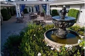Outdoor courtyard area with a stone water fountain surrounded by greenery and white flowers. In the background, there are patio tables and chairs under a pergola attached to a building.