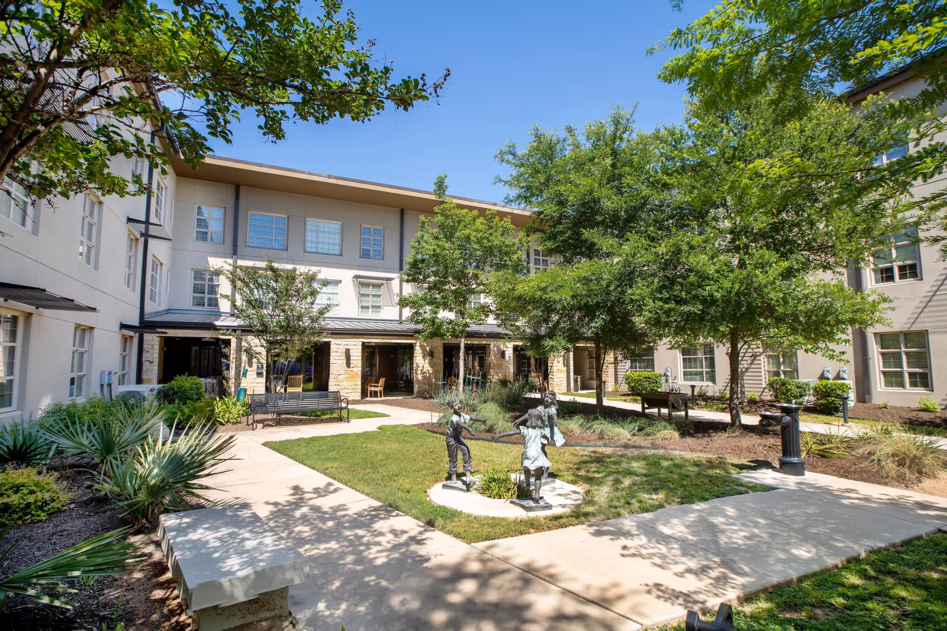 Sunny landscaped courtyard with walkways, benches, trees, small statues, and a multi-story building facade.