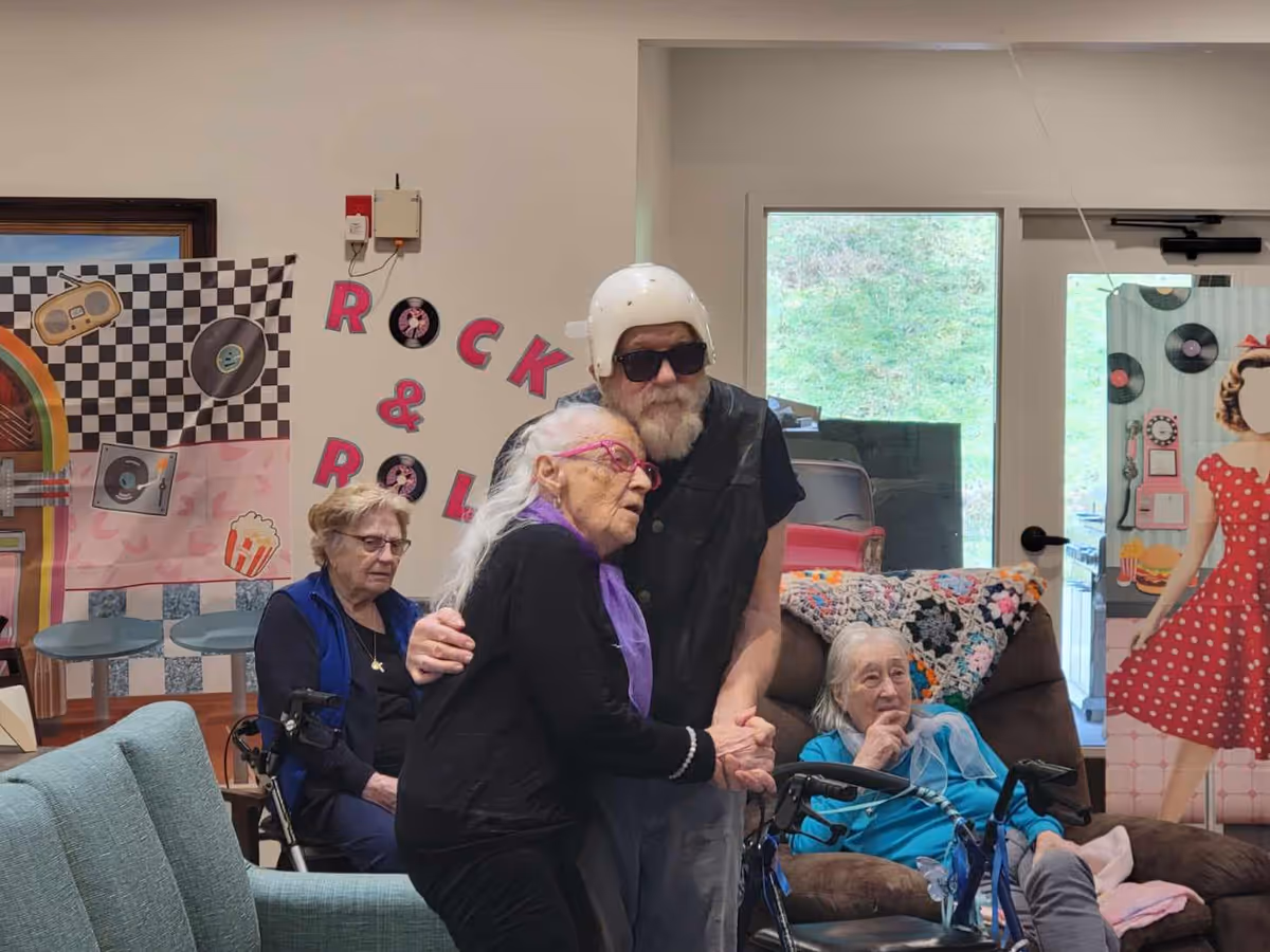 A group of elderly people in a common room decorated with a rock and roll theme. One elderly man wearing a white helmet and sunglasses is hugging an elderly woman with long white hair and pink glasses. Two other elderly women are seated nearby, one with a walker and the other on a recliner chair. The room has colorful decorations including a jukebox backdrop and a cutout of a woman in a red polka dot dress.