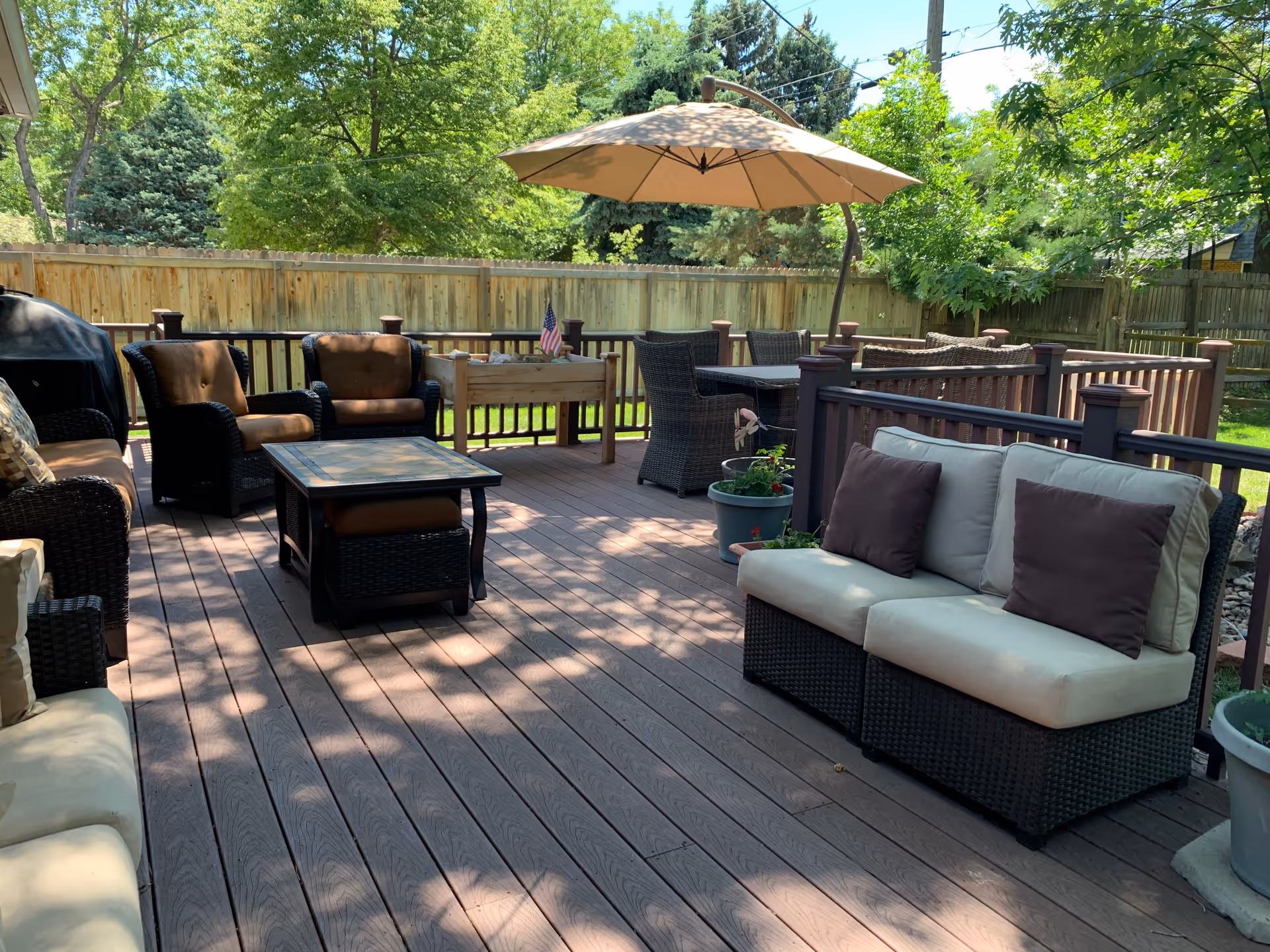 Outdoor wooden deck area with cushioned wicker seating including sofas and chairs, a table with an umbrella, potted plants, a small American flag, and a wooden fence surrounding the space with trees in the background.