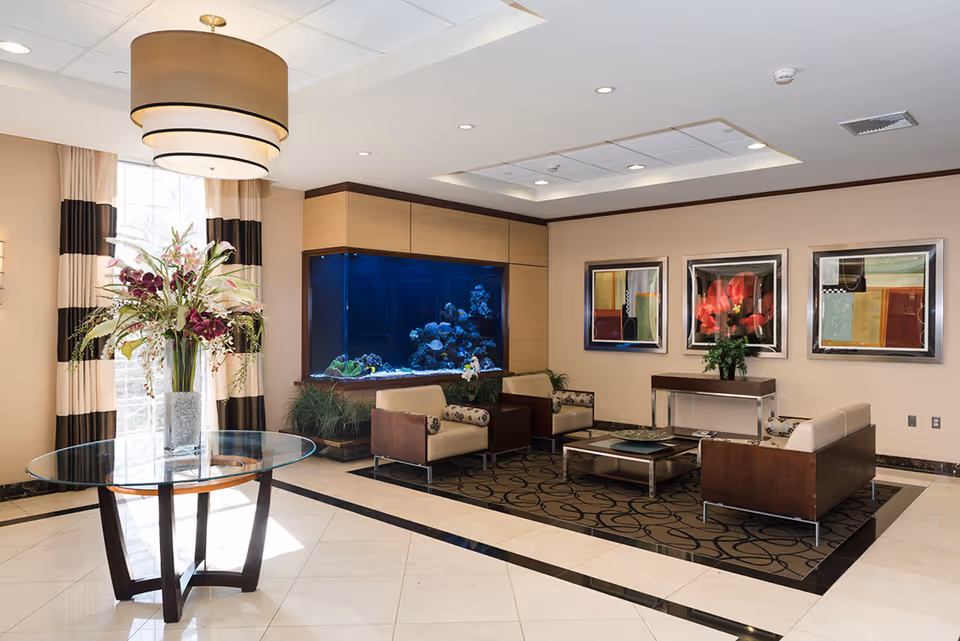 A modern and elegant seating area in a healthcare facility featuring a large built-in aquarium, four armchairs arranged around a glass coffee table on a patterned rug, a round glass table with a tall floral arrangement, large windows with striped curtains, and three colorful framed artworks on the wall.