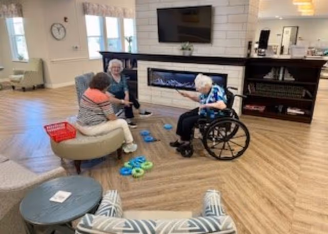 Three elderly women sitting and playing a ring toss game in a spacious, well-lit common area with wooden flooring. One woman is in a wheelchair, and the room features a modern electric fireplace, a wall-mounted TV, and comfortable seating including an ottoman and armchair. Large windows allow natural light to fill the space.