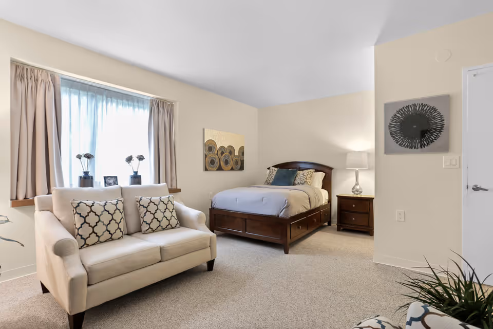 A bright and cozy bedroom featuring a wooden bed with light gray bedding and decorative pillows, a matching wooden nightstand with a lamp, a beige loveseat with patterned cushions in front of a window with beige curtains, and two pieces of abstract wall art. The room has light-colored walls and carpeted flooring.