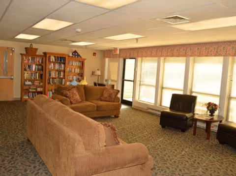A cozy living room area with two brown sofas, two dark brown armchairs, a small round table with a flower vase, and bookshelves filled with books against the wall. Large windows with blinds and a door let in natural light.