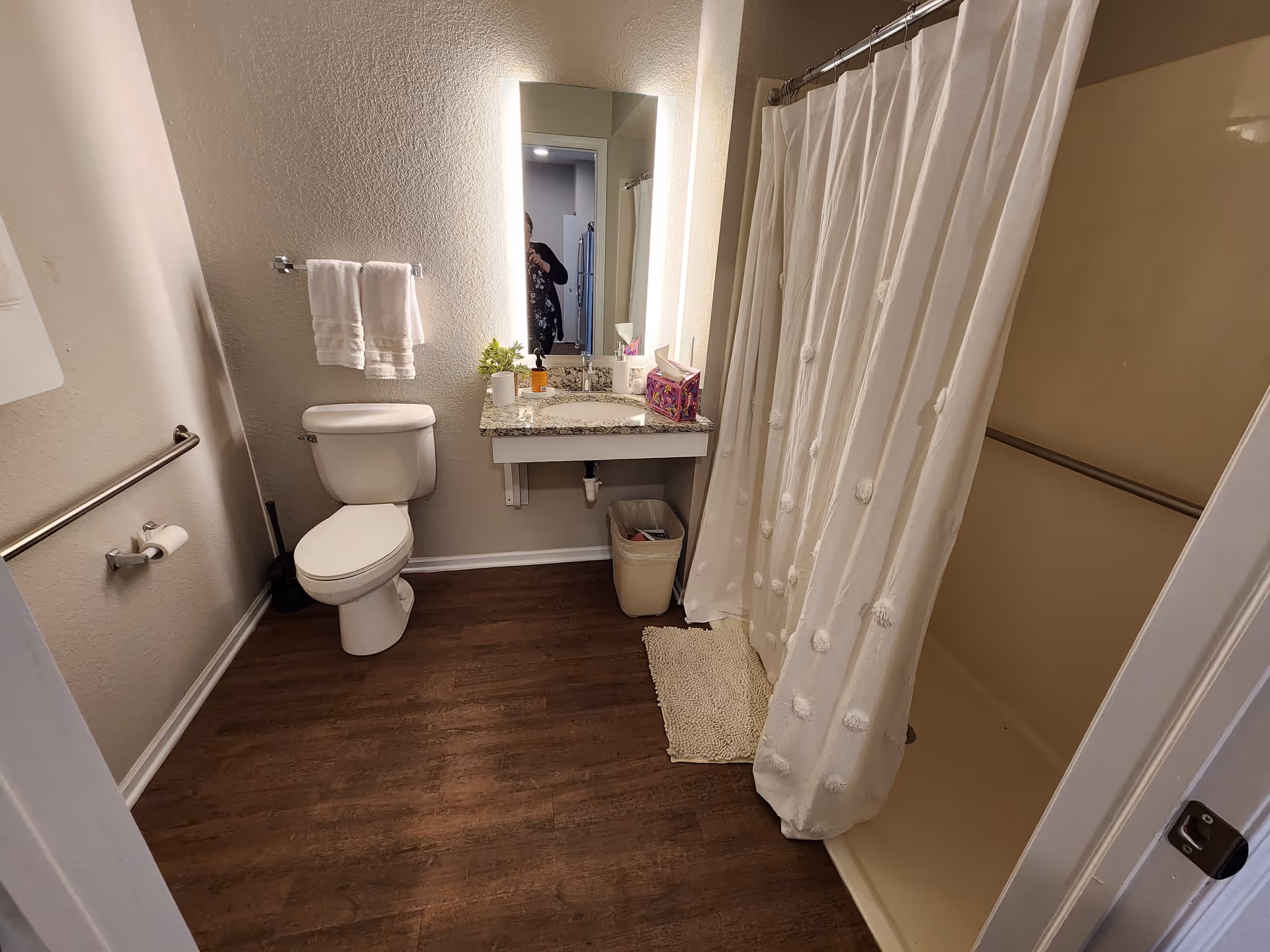 A bathroom with a toilet, a sink with a granite countertop, a mirror with built-in lighting, a shower with a white curtain, and a wooden floor. There are two white towels hanging on a towel rack above the toilet, a small plant and toiletries on the sink counter, and a trash bin underneath the sink.