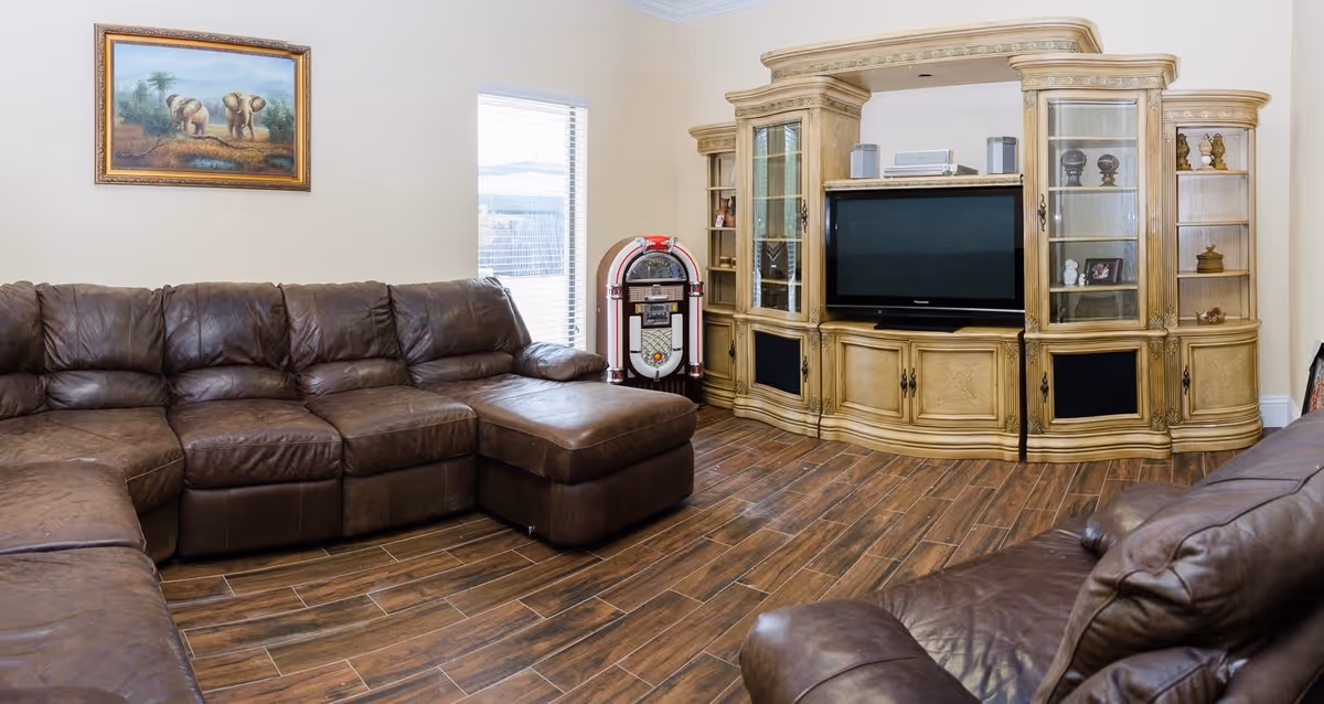 A cozy living room with brown leather sectional sofas arranged around a wooden entertainment center holding a flat-screen TV. A vintage jukebox stands next to a window with blinds, and a framed painting of two elephants hangs on the wall above the sofa. The floor is covered with wood-look tiles.