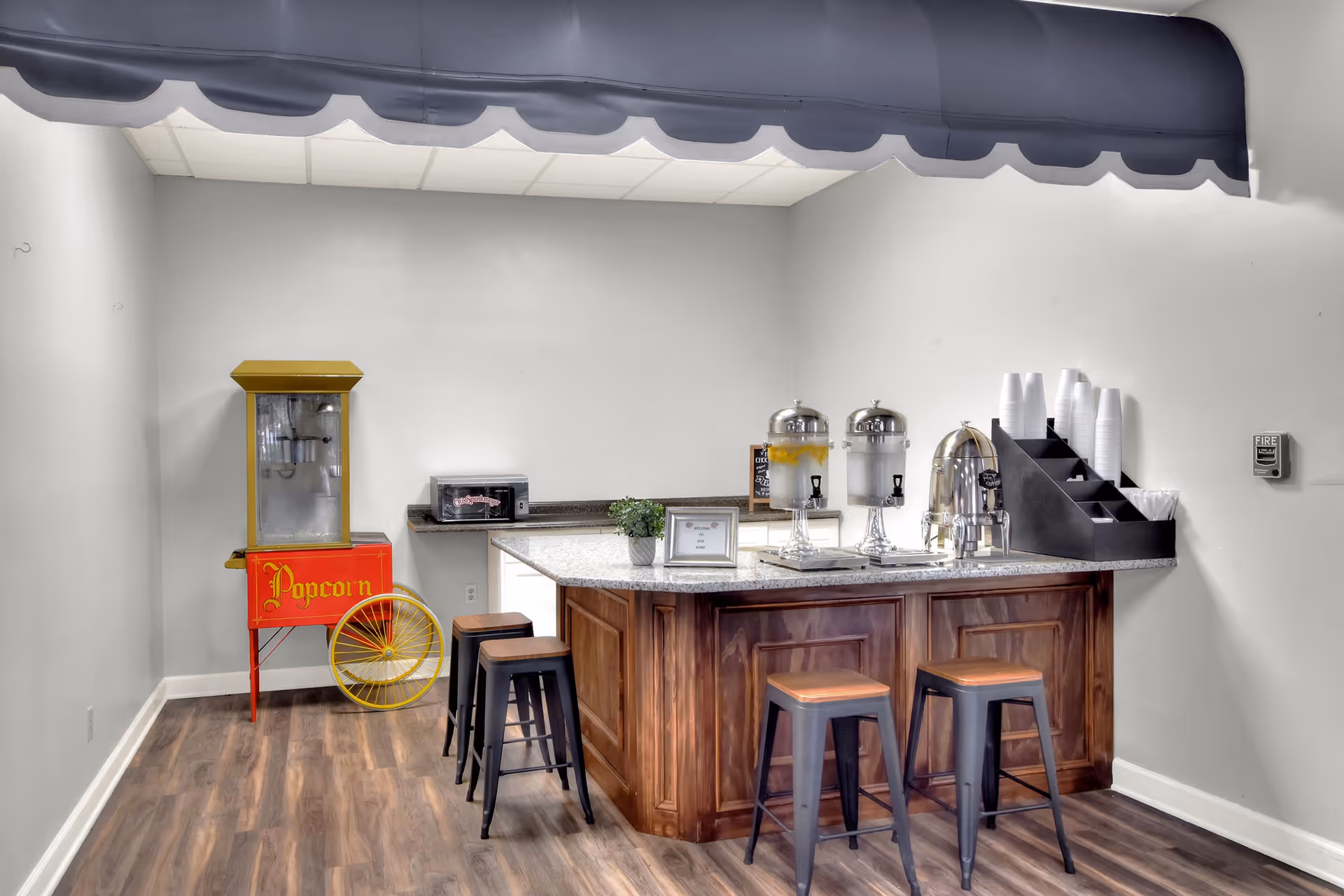 A small refreshment area with a wooden counter and four stools. On the counter are two beverage dispensers, a coffee urn, a small plant, and a framed sign. To the left, there is a vintage-style popcorn machine with a red and yellow design. The floor is wood, and the walls are light gray with a dark blue scalloped awning above the counter.