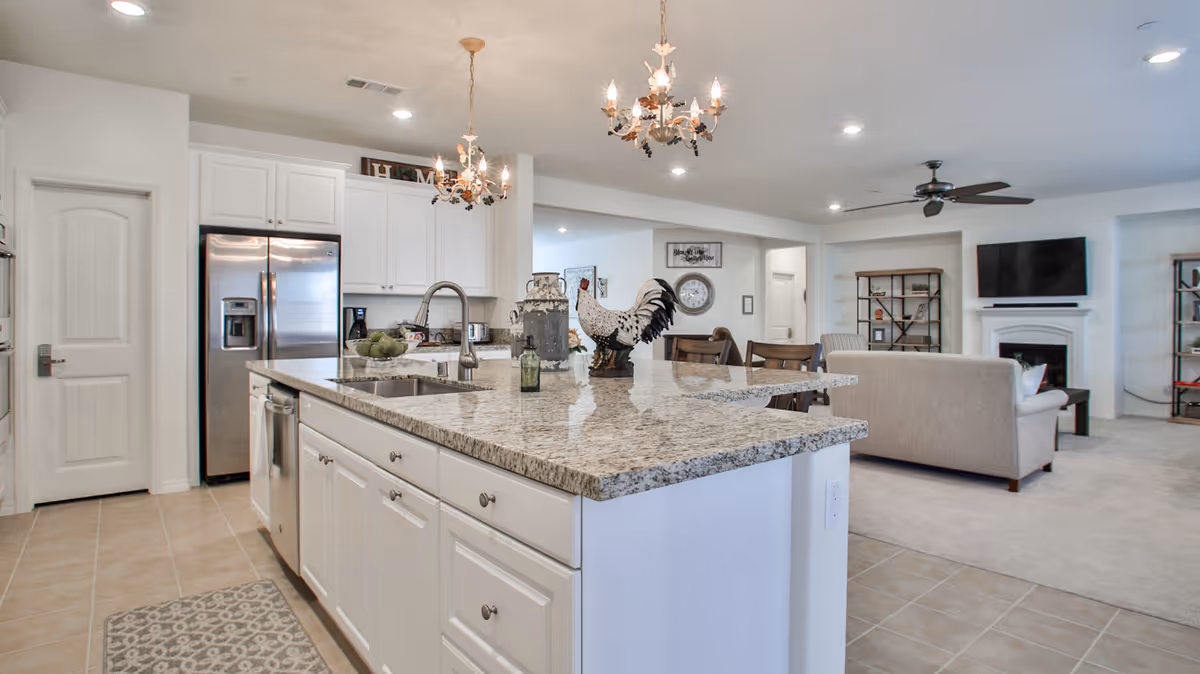 Open-plan kitchen with a large granite island, white cabinetry and stainless steel refrigerator overlooking a living area with a sofa and TV.