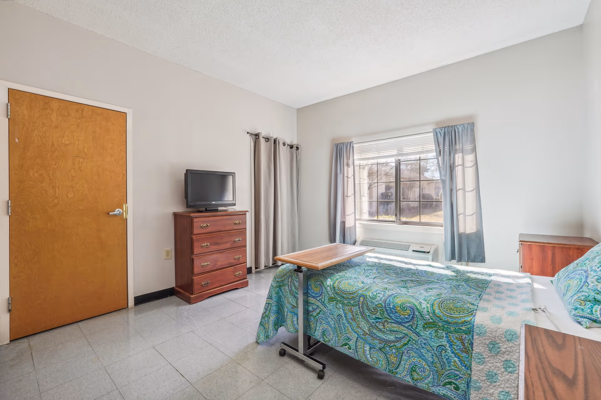 A simple bedroom with a single bed covered in a blue and green patterned quilt. There is a wooden bedside table next to the bed and a wooden dresser with a small television on top. A window with blue curtains lets in natural light, and a wooden door is visible on the left side of the room. The floor is tiled in a light gray color.