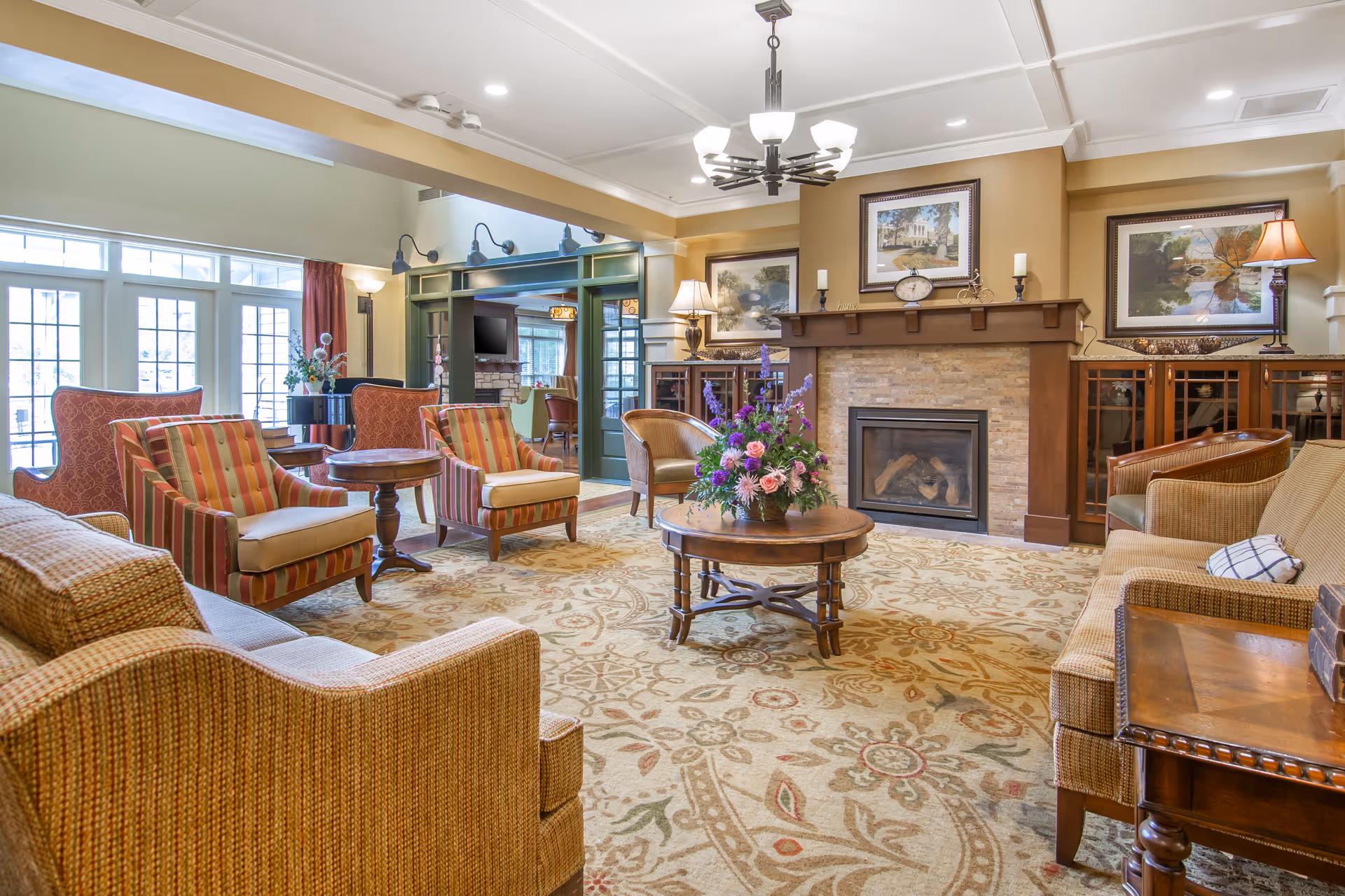 A cozy senior living facility lounge area with a fireplace, floral centerpiece on a round wooden table, multiple upholstered armchairs and sofas, framed artwork above the fireplace, and large windows letting in natural light.