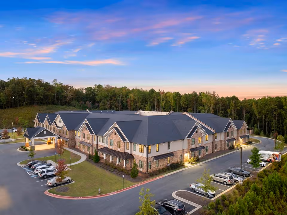 Aerial evening view of a two-story brick senior living building with a covered entrance, parking lot, and surrounding trees.
