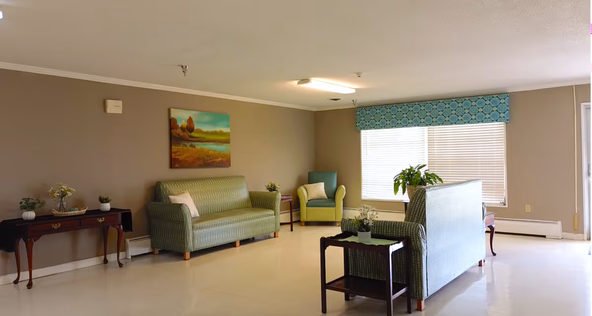A well-lit living room area with two green patterned sofas, a green armchair, a wooden side table with plants and flowers, and a large window with blinds and a blue patterned valance. The walls are painted beige and there is a landscape painting hanging above one sofa.