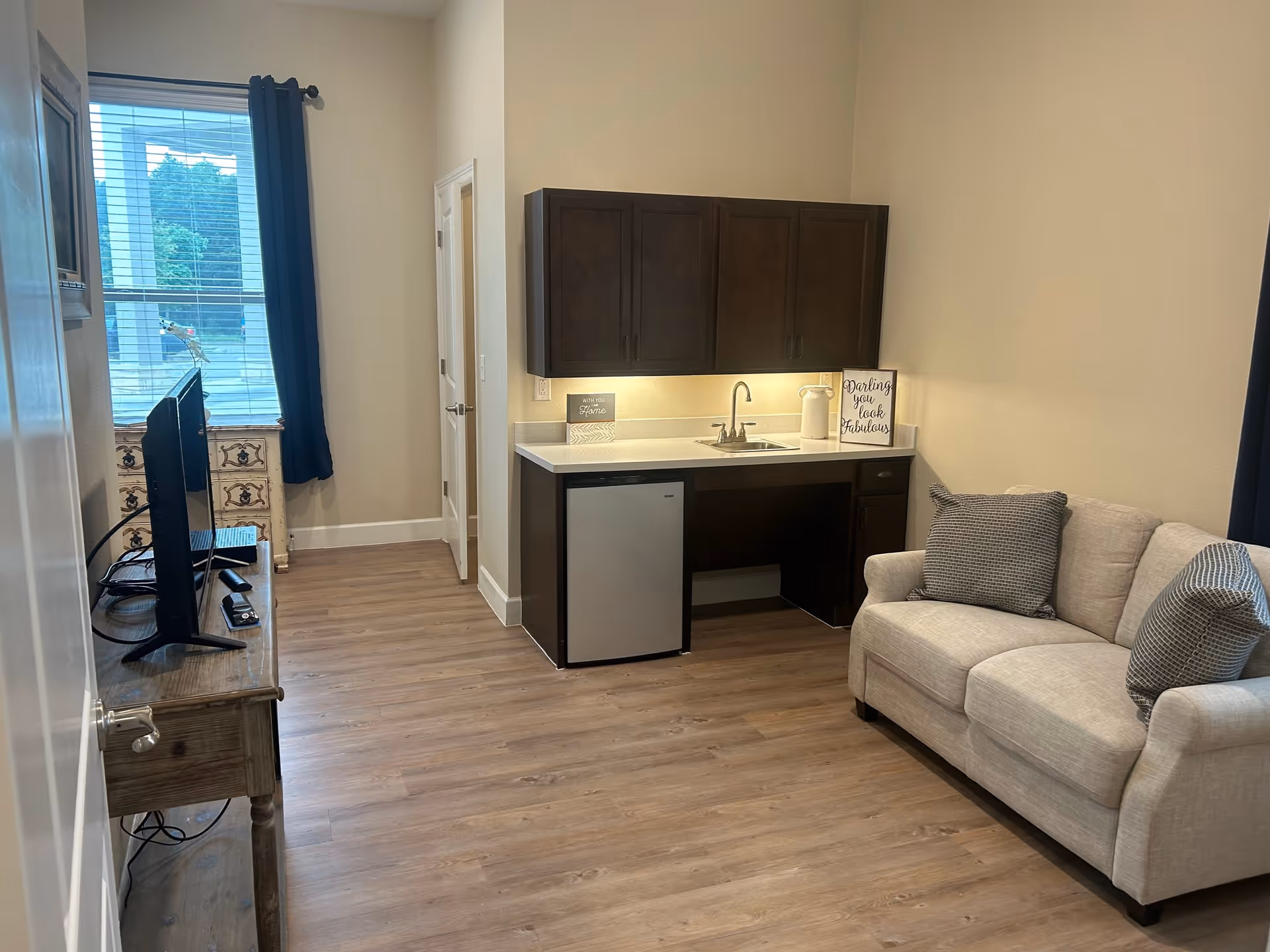 A small living area with a beige loveseat with two gray patterned pillows, a wooden TV stand with a flat-screen TV, and a kitchenette with dark brown cabinets, a small refrigerator, a sink, and countertop decor including two framed signs. There is a window with blue curtains and a door in the background.