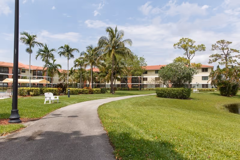A paved walkway curves through landscaped lawns and palm trees toward a multi-story senior living building with red tile roofs.