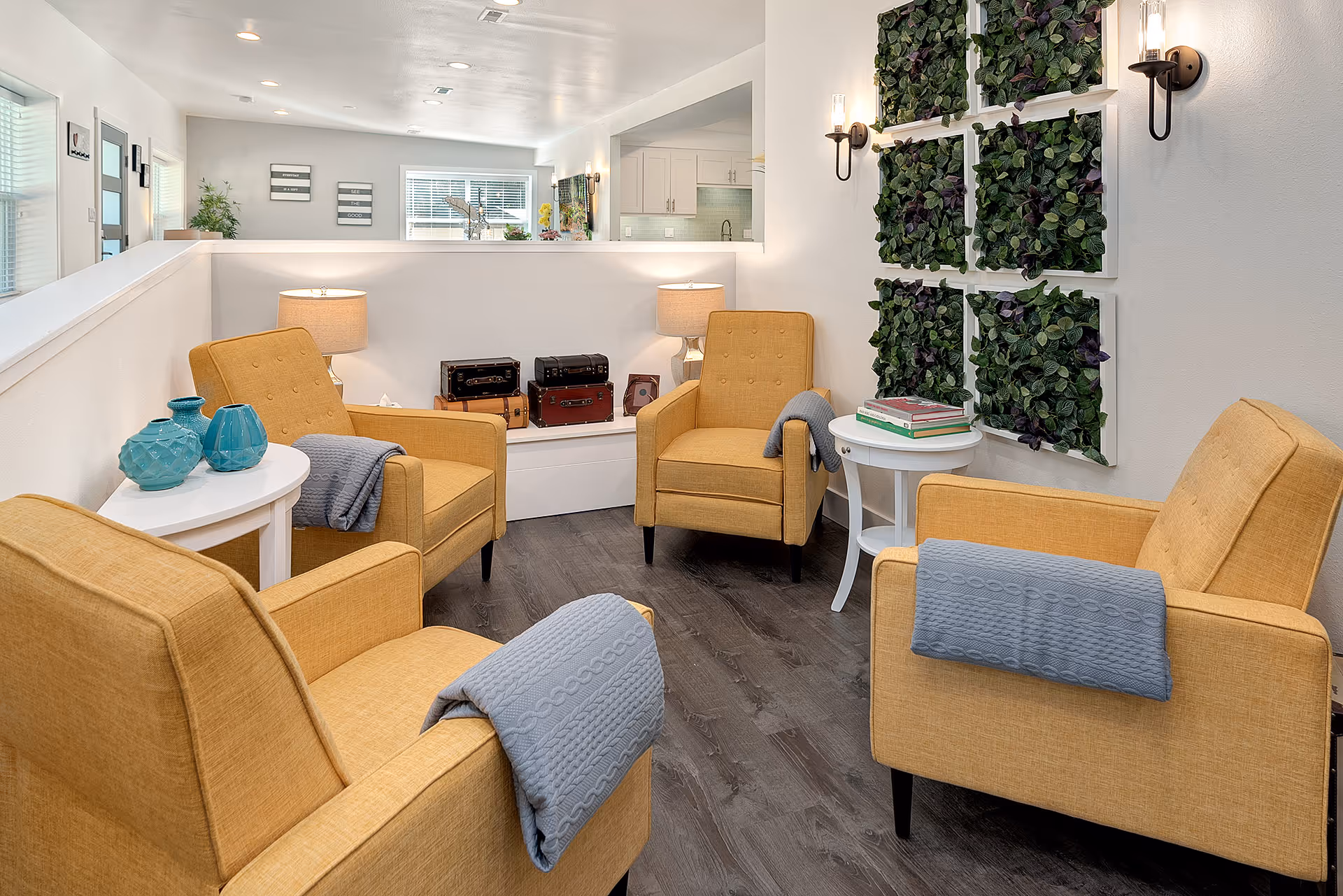 A cozy seating area with four mustard yellow armchairs arranged in a circle, each with a folded blue-gray blanket draped over the arm. Two small white side tables hold decorative items including blue vases and stacked books. The room has dark wood flooring, white walls, and a decorative green plant wall art. In the background, a kitchen area is visible with white cabinetry and a window letting in natural light.