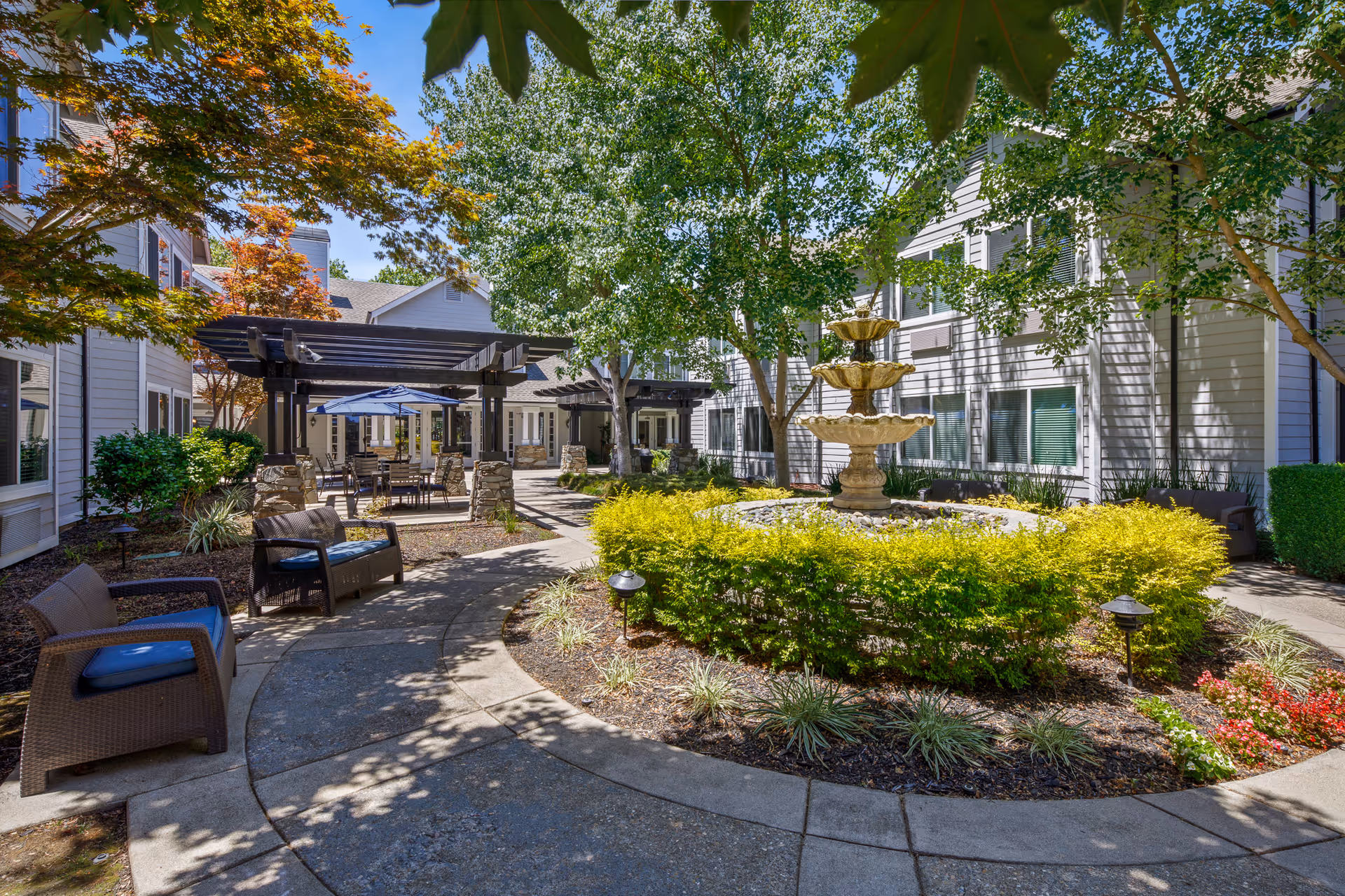 Sunlit courtyard with a tiered fountain, seating areas, a pergola and surrounding residential building.