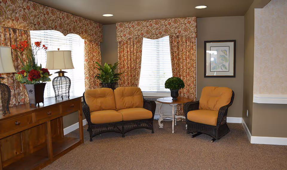 A cozy sitting area with two wicker chairs and a loveseat with mustard yellow cushions, a small round wooden table with a potted plant, floral patterned curtains on two windows, a wooden sideboard with two lamps and a flower arrangement, and a framed botanical print on the wall.