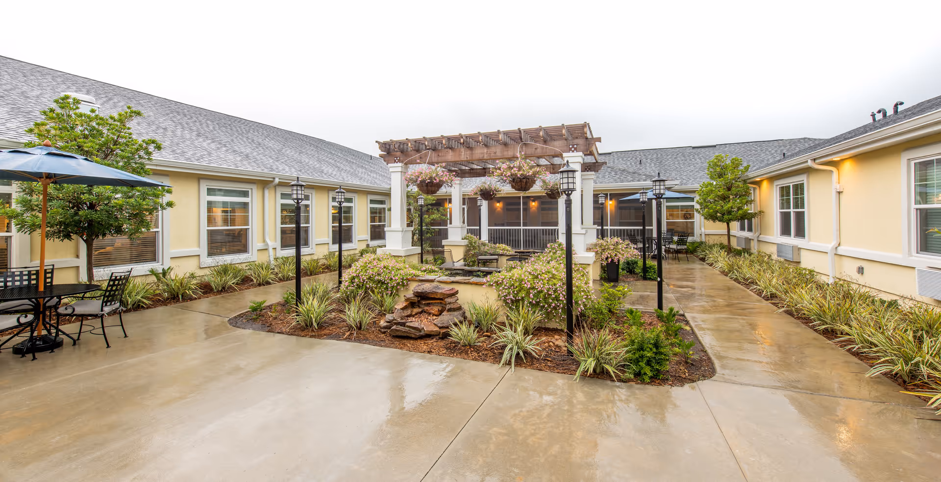 Outdoor courtyard area at The Addison of Oakleaf featuring a pergola with hanging flower baskets, a small rock water feature, surrounding plants, patio tables with umbrellas, and a wet concrete walkway between yellow buildings with multiple windows.