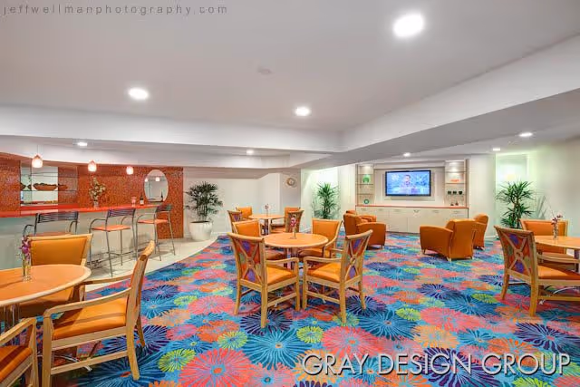 A brightly lit common area with colorful floral carpet, several round wooden tables with orange cushioned chairs, a bar area with high stools and pendant lights, and a wall-mounted television surrounded by built-in shelves and plants.