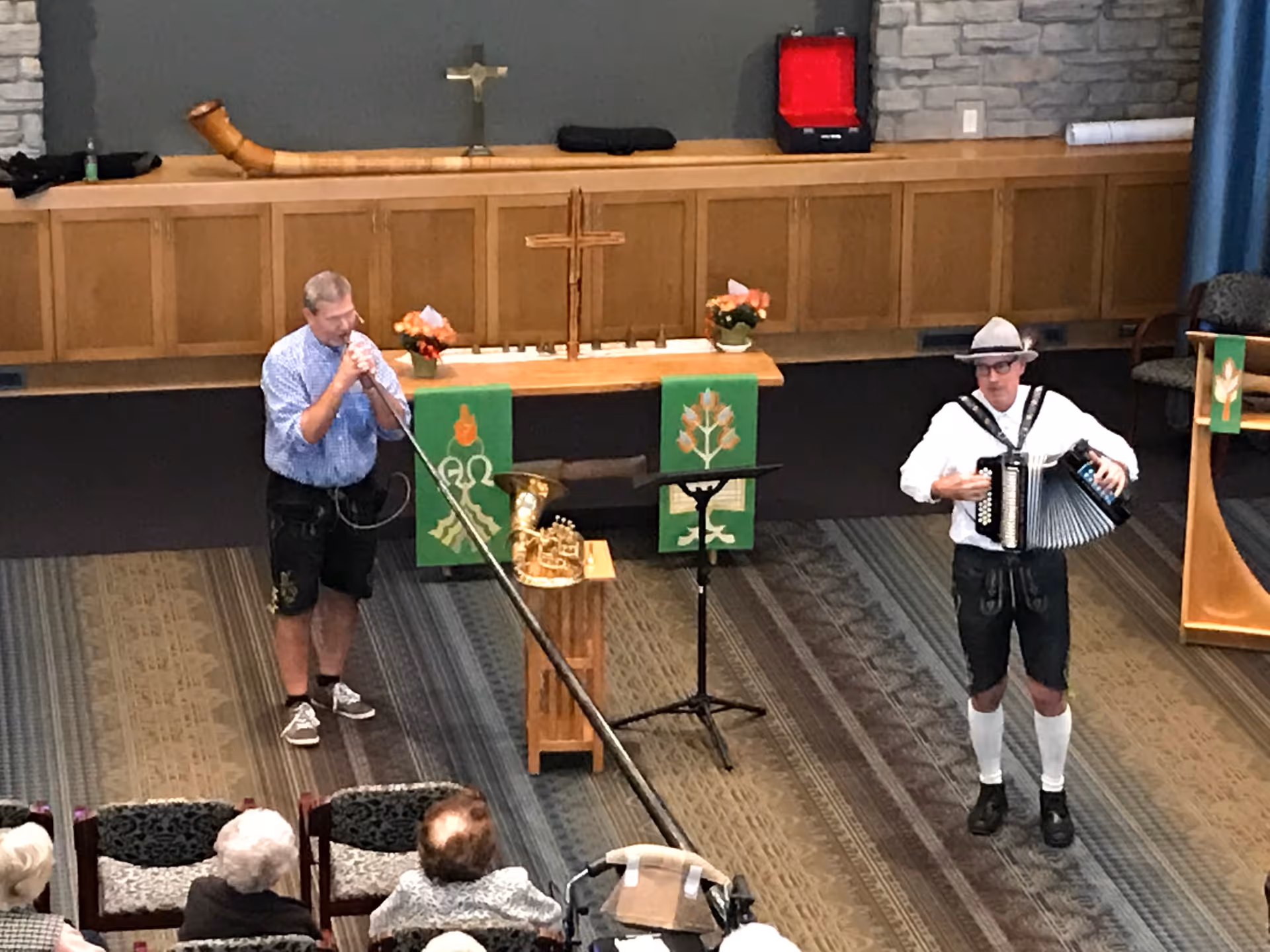 Two men performing music in a church or chapel setting. One man is playing a long alphorn, and the other is playing an accordion. There are a few seated elderly audience members watching the performance. The background shows a wooden altar with a cross, flowers, and green cloth banners with religious symbols.