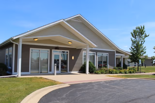 Exterior view of a single-story building with gray siding and white trim, featuring a covered entrance with two ceiling lights, large windows, and a paved driveway with a curb. There is a small landscaped area with bushes and a tree near the entrance under a clear blue sky.