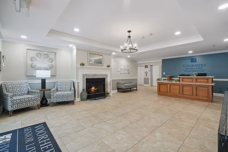 Reception area of Voorhees Senior Living featuring a wooden front desk with two chairs behind it, a blue accent wall with the facility's name and logo, a fireplace with a decorative mantle, two patterned armchairs with a small round table and lamp between them, a bench along the wall, and tiled flooring.
