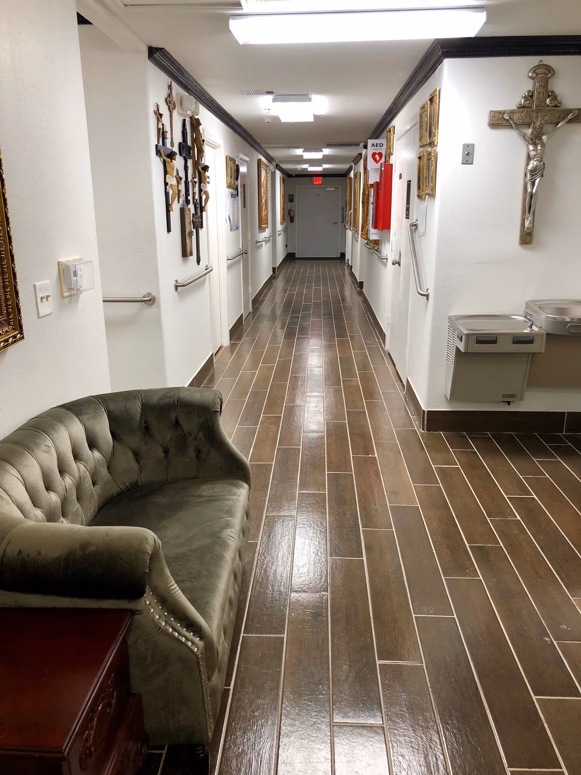 A long hallway in an assisted living facility with brown tiled flooring and white walls. The walls are decorated with multiple religious crosses and framed pictures. On the left side, there is a green tufted sofa next to a wooden side table. On the right side, there are two water fountains and a red AED box mounted on the wall. The hallway is well-lit with ceiling lights and has handrails on both sides.
