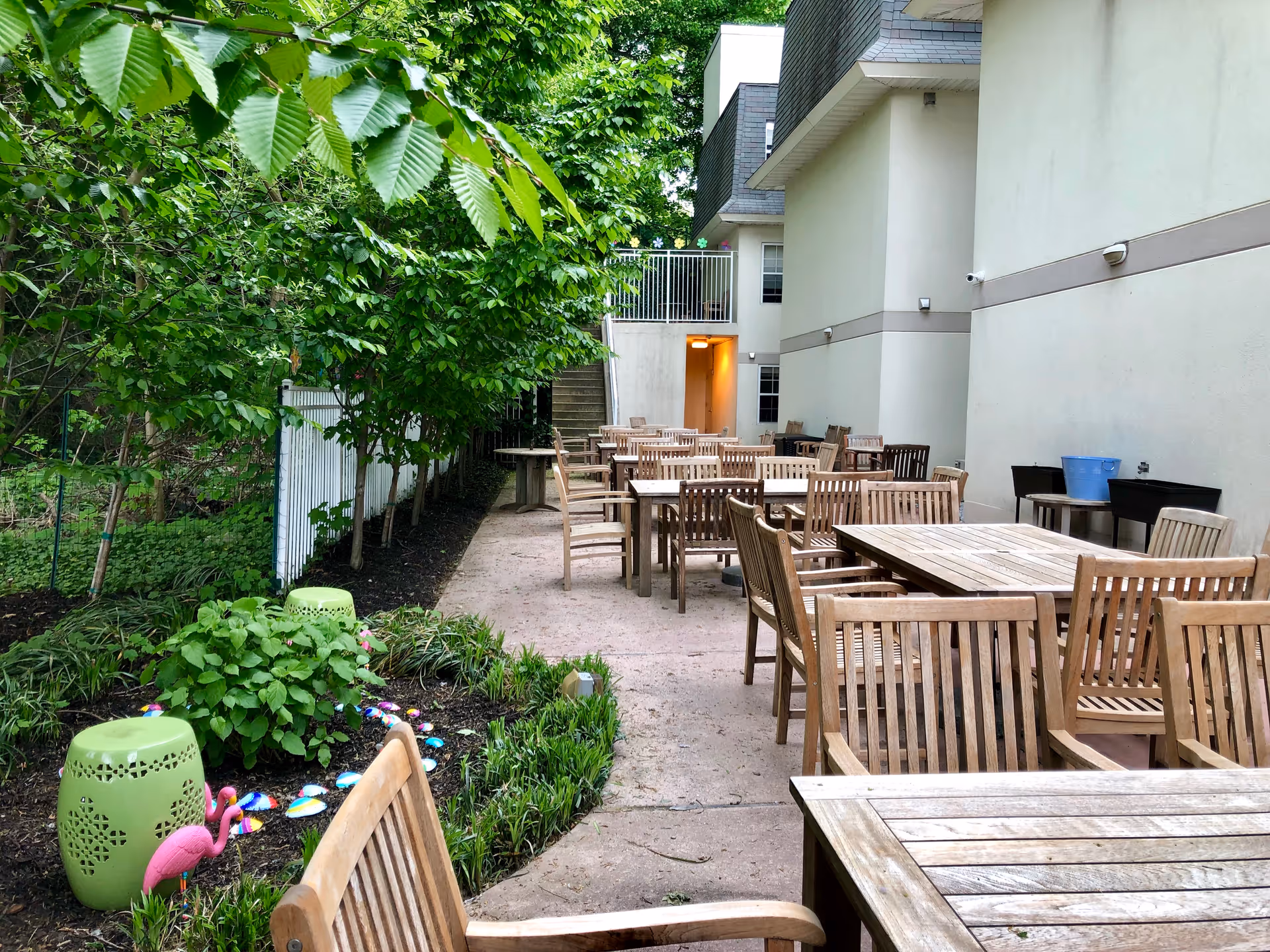 Outdoor patio with multiple wooden tables and chairs next to a building and a small garden area.