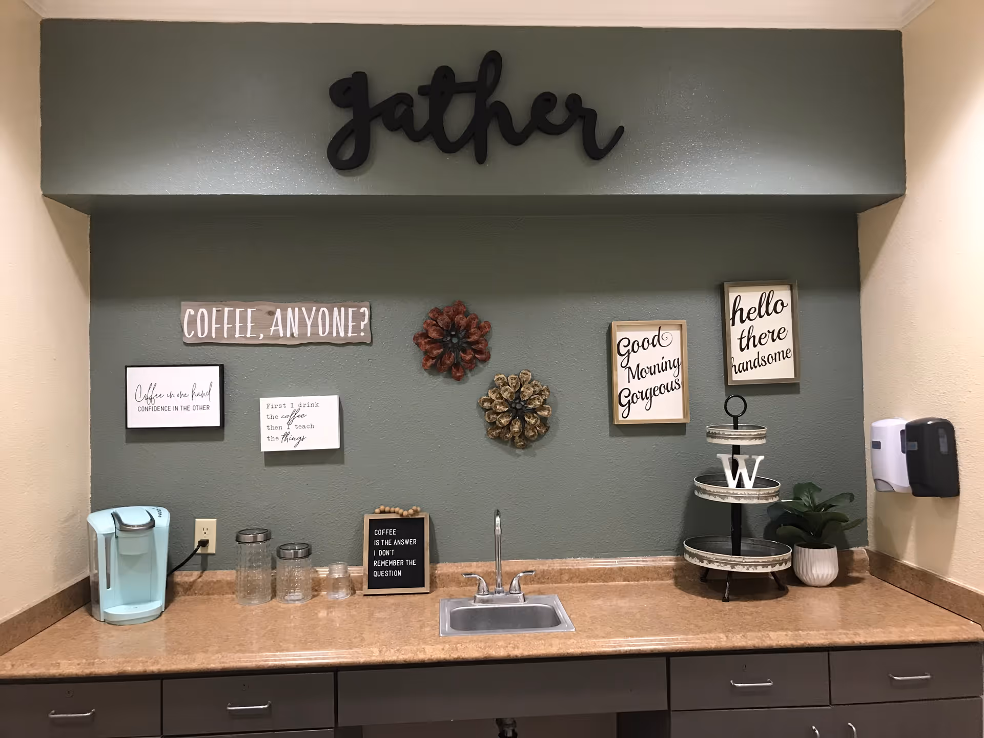 A small kitchenette area with a countertop, a sink, a light blue coffee maker, glass jars, and decorative signs on the wall. The wall is painted green with the word 'gather' in large black letters above. Various framed signs with coffee-related and welcoming messages are displayed, along with decorative pinecones and a tiered tray with a plant and a letter 'W'.