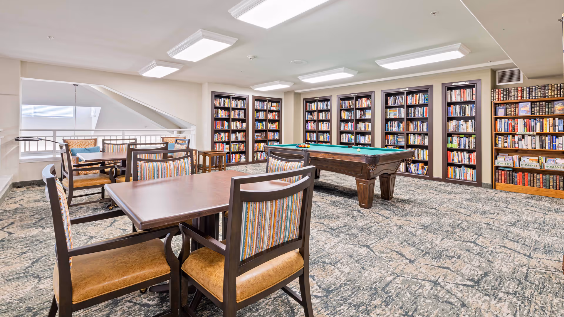 A bright and spacious common room featuring a pool table in the center and multiple bookshelves filled with books along the walls. There are several wooden tables with cushioned chairs arranged around the room, creating a comfortable space for reading, playing games, or socializing.