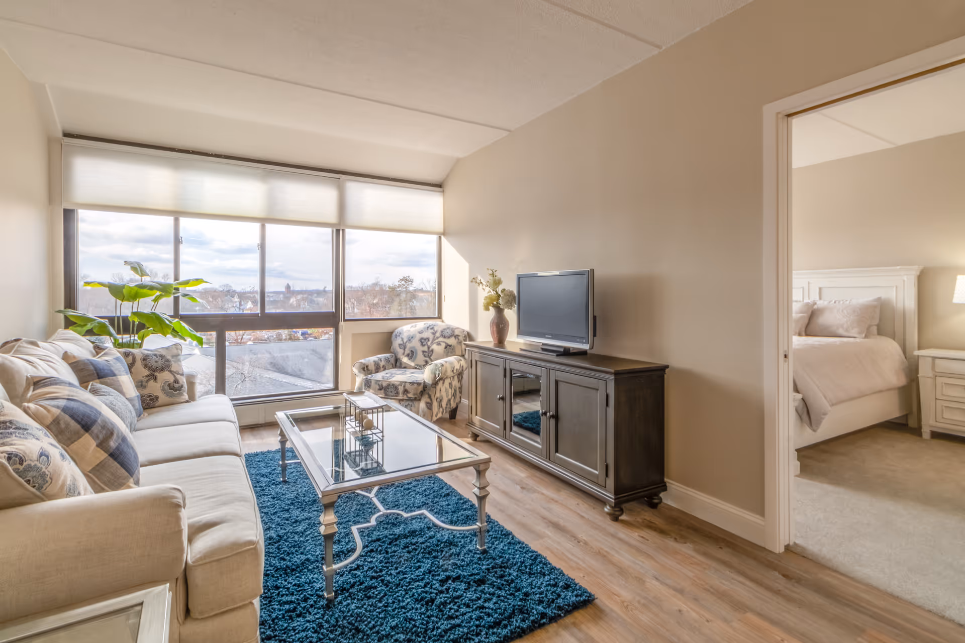 A bright living room with a large window letting in natural light. The room features a beige sofa with patterned cushions, a glass coffee table on a blue rug, a floral armchair, and a dark wooden TV stand with a flat-screen TV and a vase with flowers. Through an open doorway, a bedroom with a white bed and nightstand with a lamp is visible.