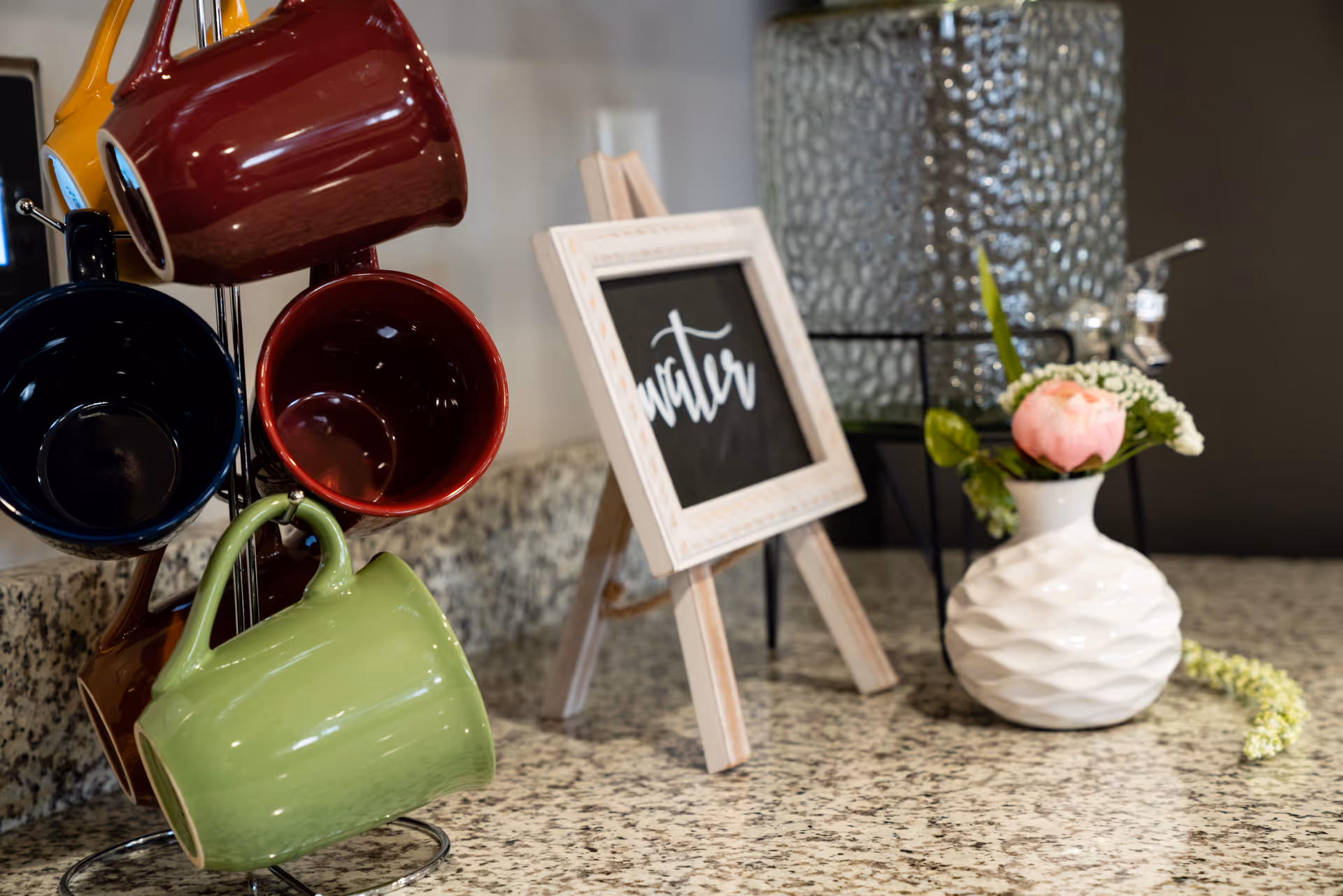 Colorful coffee mugs on a rack next to a small chalkboard sign and a vase with a pink flower on a granite countertop.