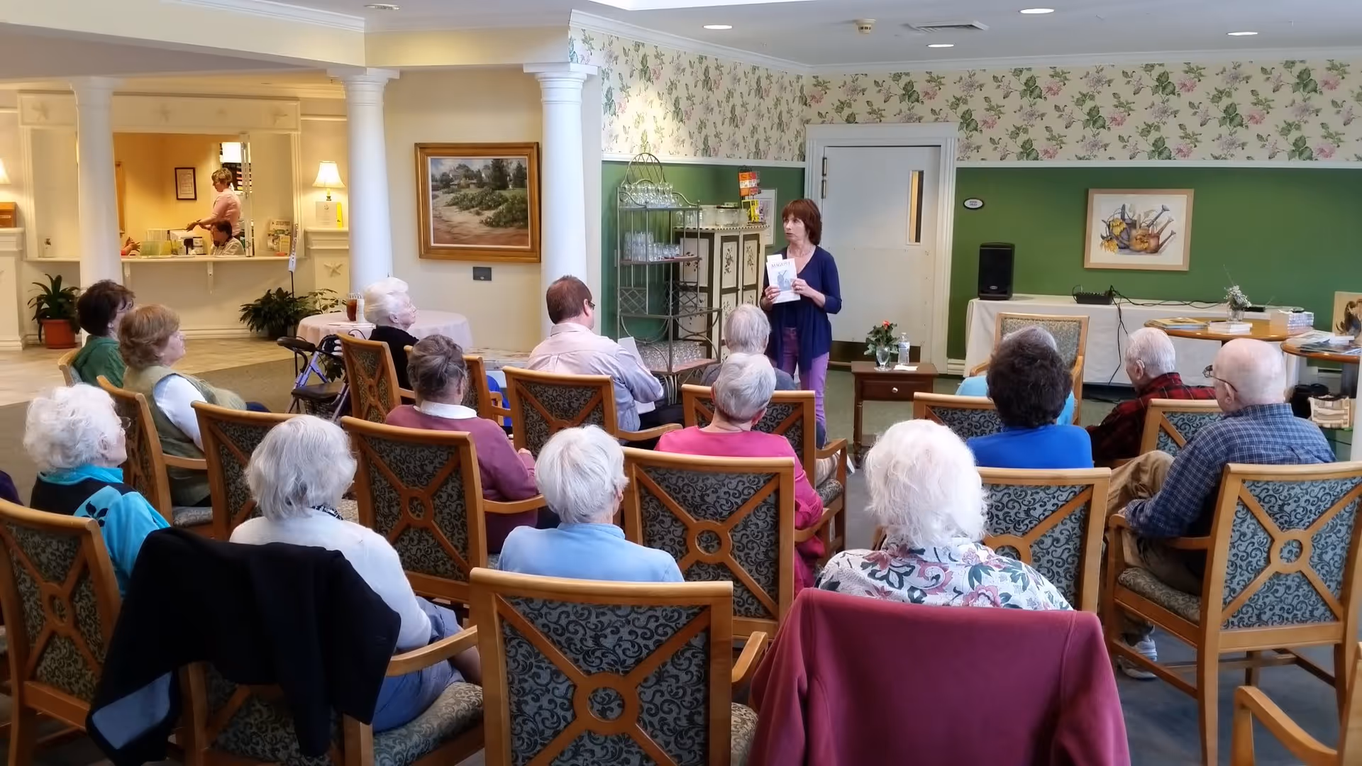 A group of elderly people seated in a room facing a woman who is standing and holding a book, possibly giving a presentation or reading. The room has floral wallpaper, green walls, and decorative columns. There is a reception area visible in the background with two staff members.