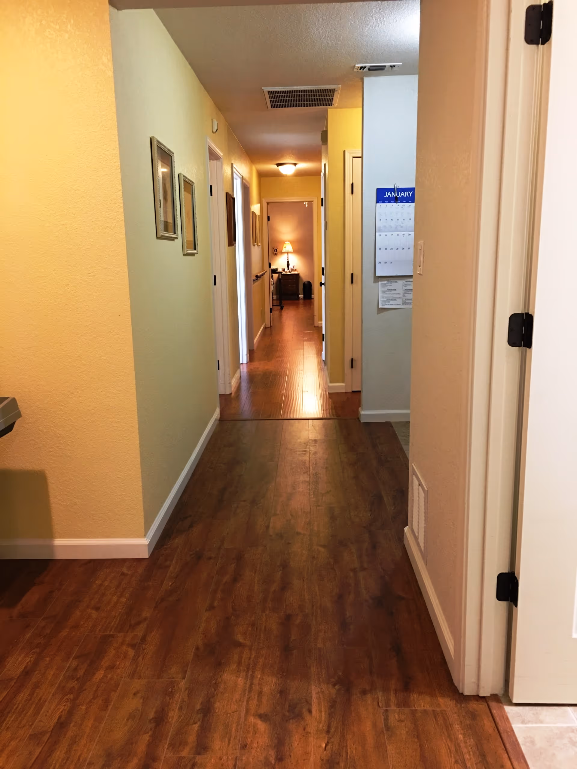 A long hallway with wooden flooring and light yellow walls. Several doorways line the hallway on both sides, and framed pictures hang on the left wall. At the end of the hallway, a small table with a lamp is visible, casting a warm light. A calendar is hanging on the right wall near the foreground.