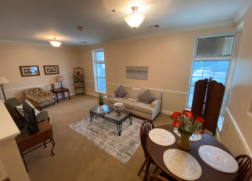 A cozy living room with beige walls and carpet. The room features a beige sofa with three cushions, a glass coffee table on a patterned rug, an armchair with floral upholstery, a wooden side table with a lamp, and a small TV on a stand. There is a round wooden dining table with four chairs and a vase of red flowers. Two tall windows with blinds allow natural light into the room, and a decorative folding screen is placed near the windows.