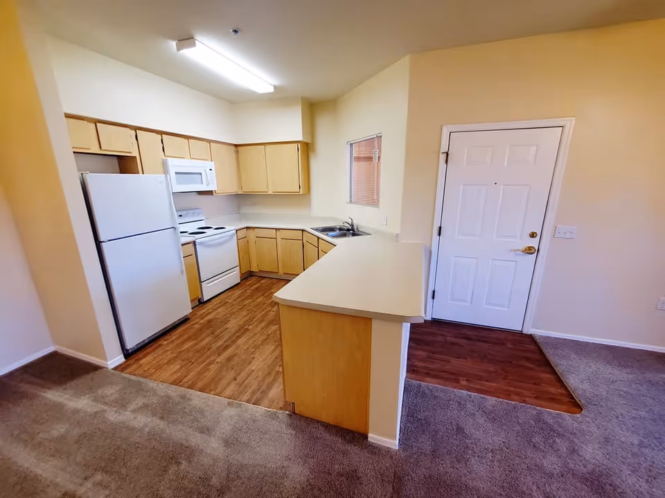 Interior view of a kitchen area in a senior living facility with light wood cabinets, white refrigerator, stove, microwave, and a sink. The kitchen has wood flooring and a countertop that extends into a small bar area. Adjacent to the kitchen is a white door and carpeted flooring in the living area.