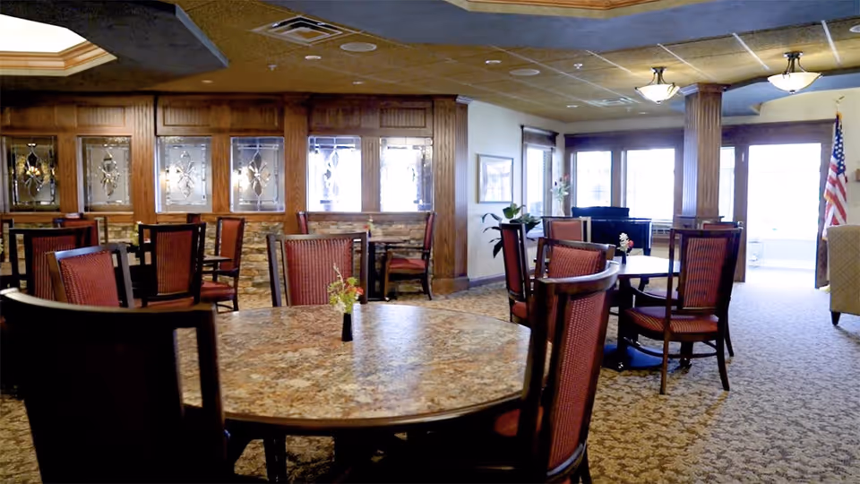 A dining room with multiple round tables and chairs featuring red upholstery. The room has carpeted floors, decorative wooden paneling with glass inserts, and large windows letting in natural light. An American flag is visible near the entrance.