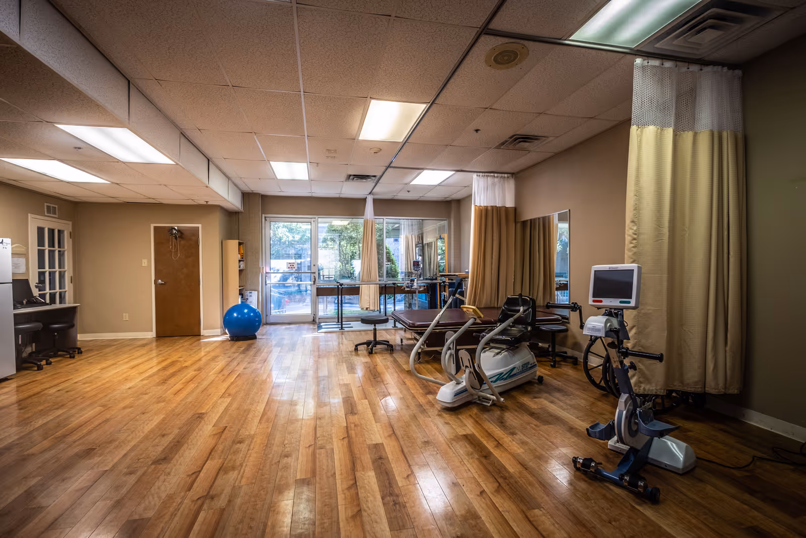 A spacious room with wooden flooring featuring exercise equipment including a stationary bike and a recumbent bike, a blue exercise ball, a therapy table, stools, and a desk with a computer. Large windows and glass doors at the back provide natural light and a view of an outdoor area. Beige curtains hang from the ceiling, partially enclosing the therapy area.