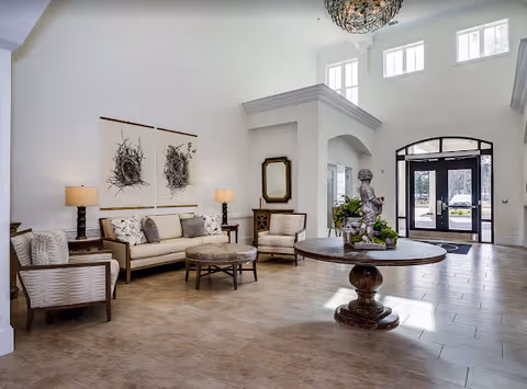A spacious and bright living room area with high ceilings and large windows. The room features a beige sofa with patterned cushions, two matching armchairs, a round ottoman, and two table lamps on side tables. A round wooden table with a decorative statue and plants is in the center. The entrance door with glass panels is visible in the background.