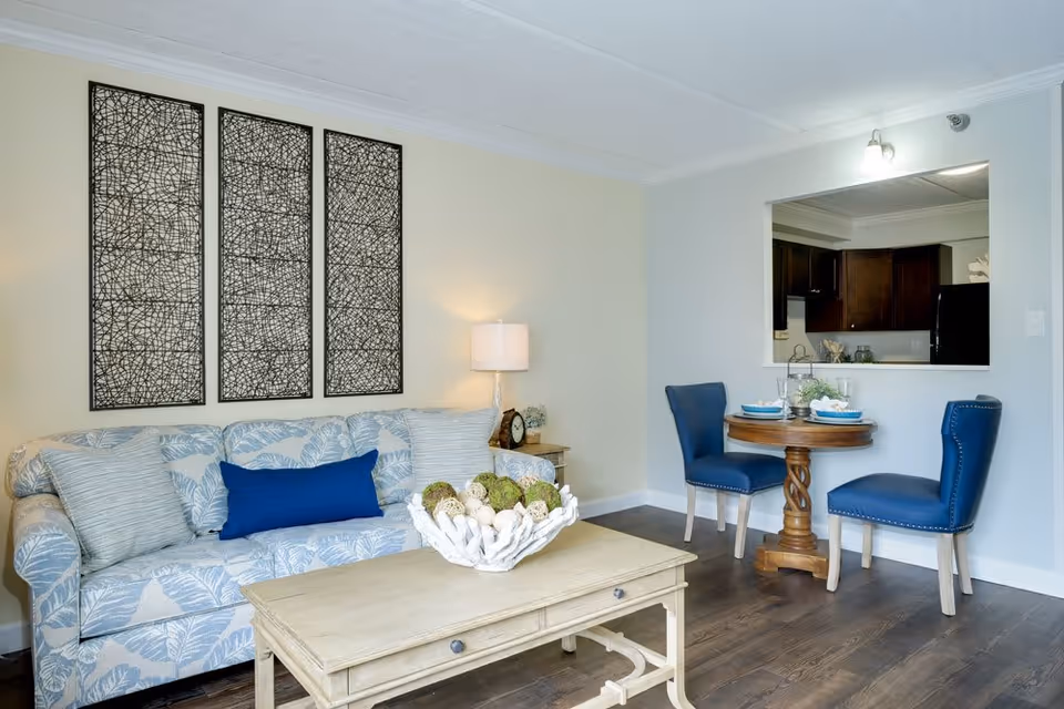 A cozy living area with a light blue patterned sofa adorned with cushions, a wooden coffee table with decorative items, a side table with a lamp and clock, and a small round dining table with two blue upholstered chairs. The dining area is adjacent to a kitchen visible through a pass-through window.