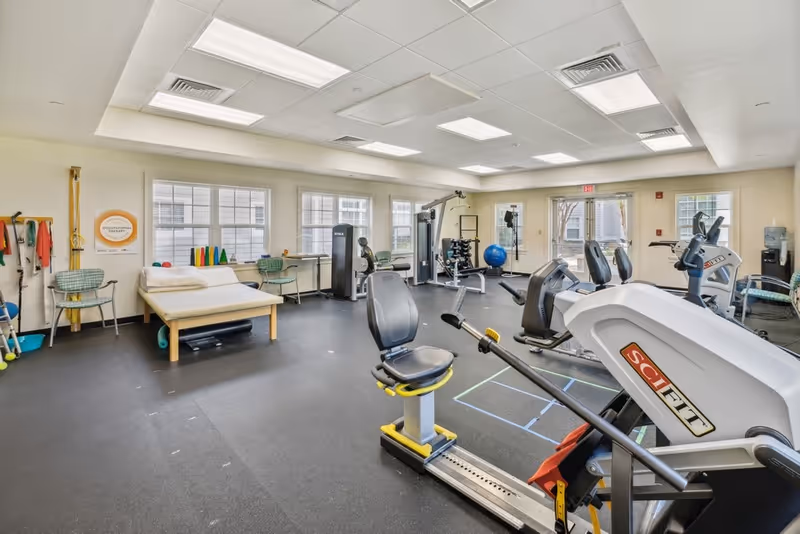A spacious fitness room in a senior living facility with various exercise equipment including stationary bikes, weight machines, and a therapy table. The room has large windows letting in natural light, chairs along the walls, and exercise accessories like resistance bands and exercise balls.