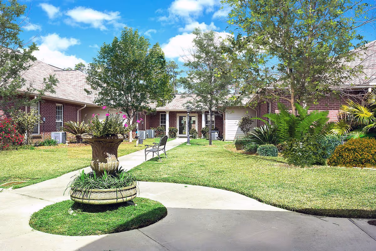Outdoor courtyard area of a senior living facility with a circular flower planter in the center, green grass, trees, benches, and a brick building with multiple windows and doors under a blue sky with some clouds.
