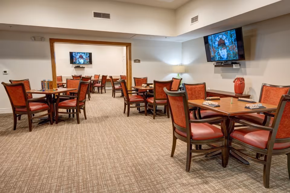 A spacious common area with multiple wooden tables and red cushioned chairs arranged for group activities. Two wall-mounted flat-screen TVs are visible, one in the foreground and one in the adjoining room. The room is carpeted and well-lit with a lamp in the corner and neutral-colored walls.