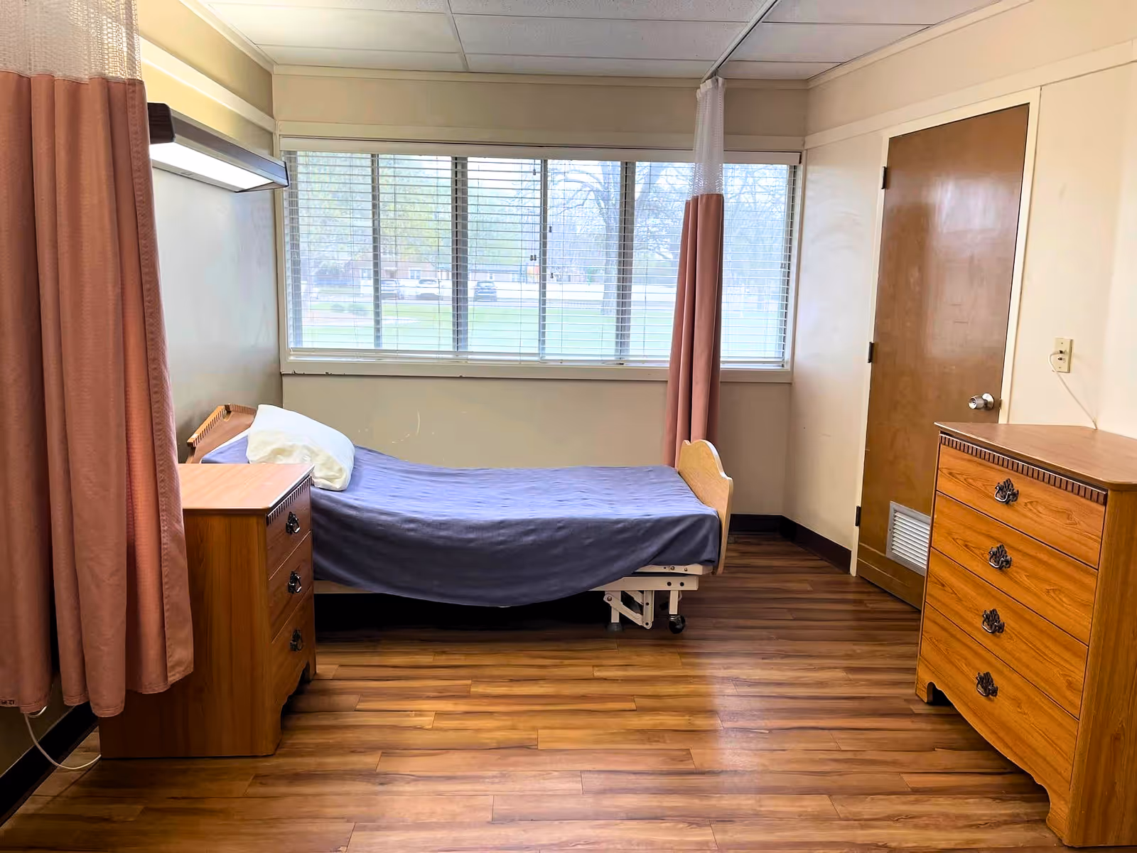 A simple nursing home bedroom with a single bed covered in a blue blanket and a white pillow. The room has wooden flooring, a wooden dresser with four drawers on the right, and a smaller wooden nightstand with three drawers on the left. There are large windows with blinds letting in natural light, and pink privacy curtains partially drawn around the bed. A wooden door is visible on the right side of the room.